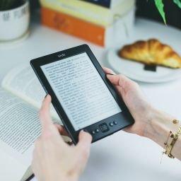 mani che tengono un tablet con ebook e sfondo di libri, tazze e un croissant